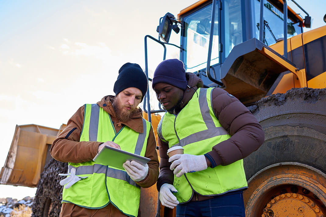 two construction workers look at a tablet while standing in front of a bulldozer