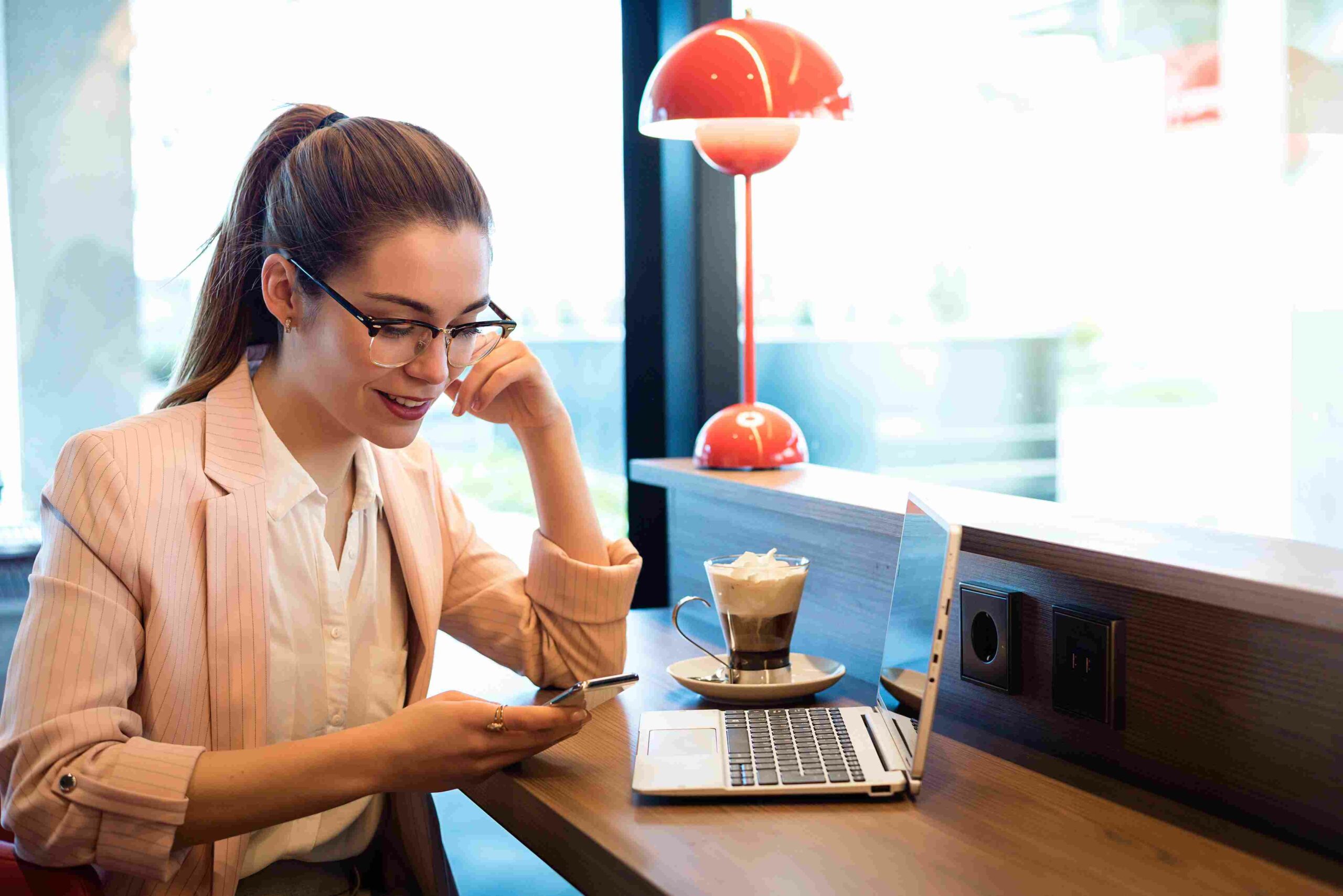 Young woman having coffee, scrolling through phone with laptop near her