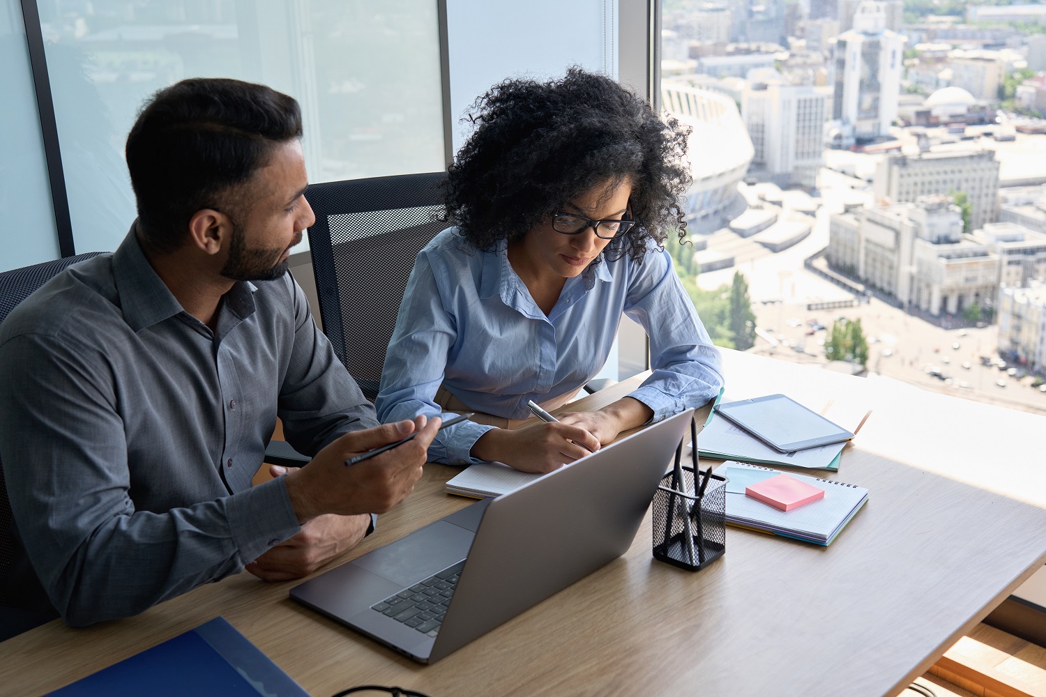 Multiethnic coworkers. Male Indian mentor and woman African American intern sitting at desk with laptop working together discussing project. Woman is taking notes.