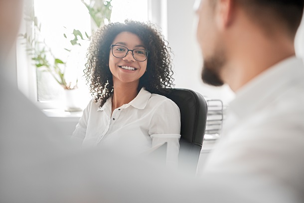 Smiling woman with curly hair sitting in black chair talking to man and woman in white shirts across the table in foreground who are blurry