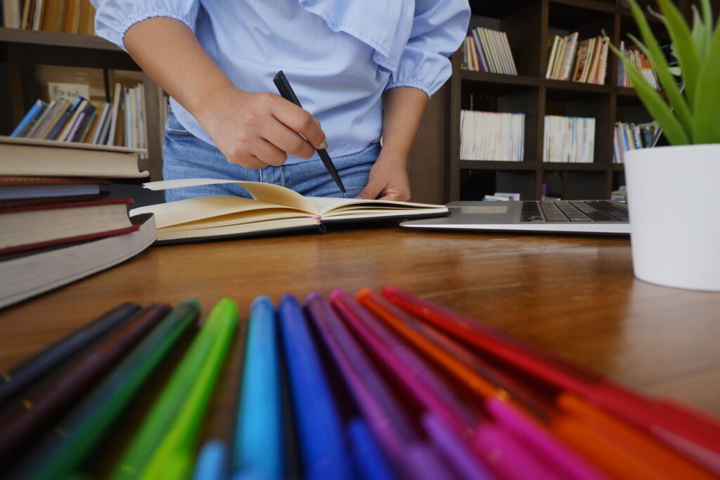 Close-up of woman's hand writing with a pen in a book. Pens of all colours on desk in front of her.