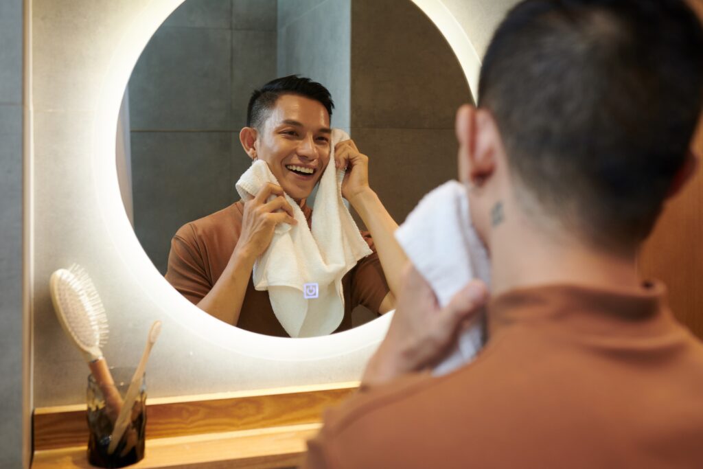 Cheerful young man wiping face with soft towel after washing it in the morning