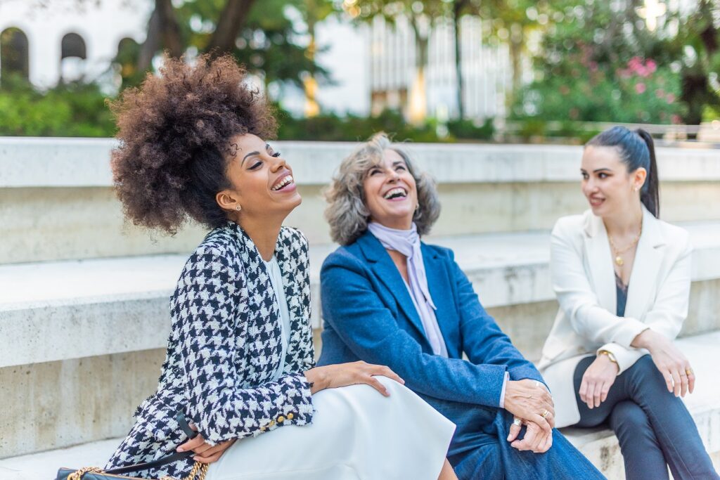 Three women in blazers laughing together - one with curly hair and houndstooth white/black blazer, one with grey hair + blue blazer + purple scarf, one with black hair in ponytail in white blazer