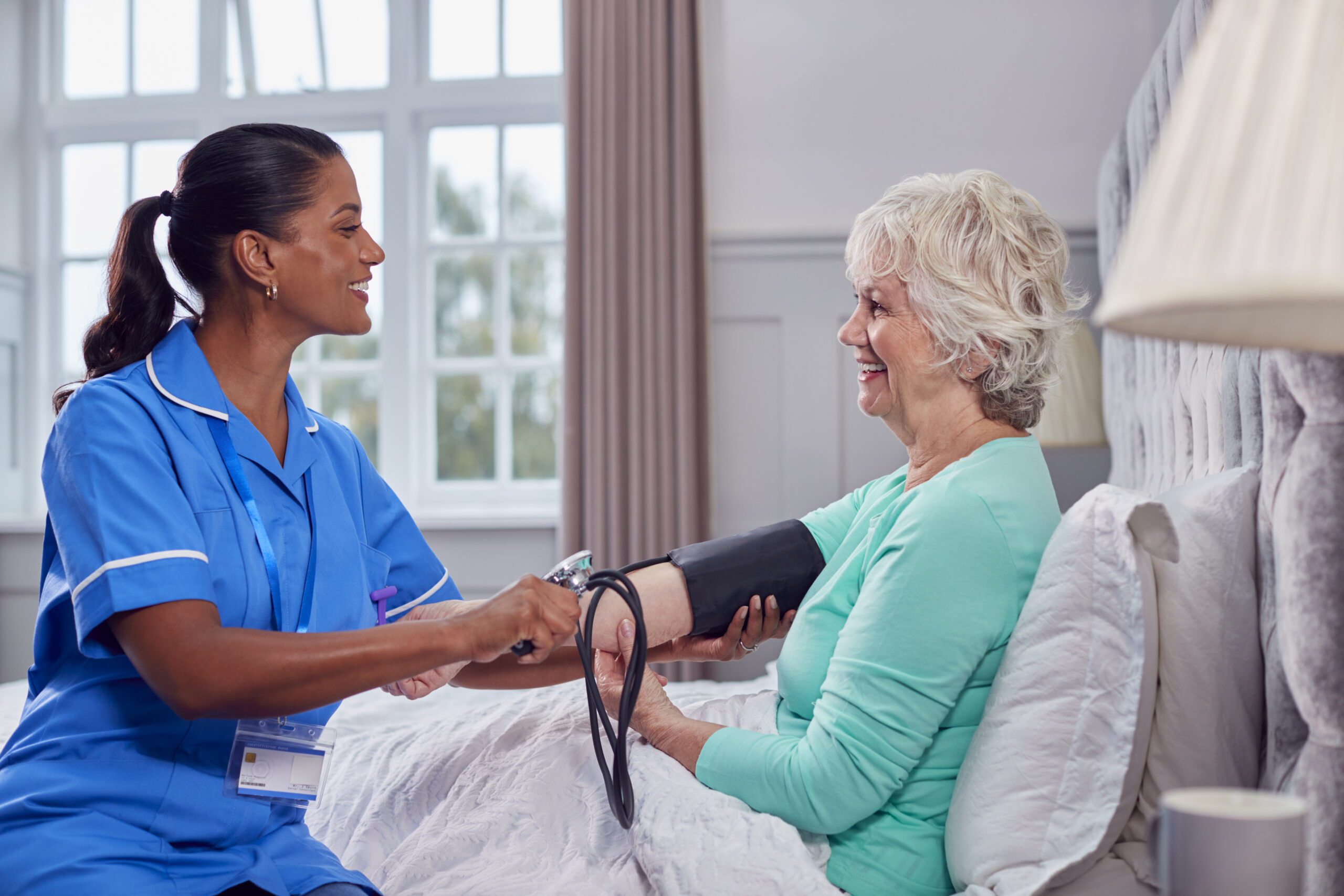 Senior Woman At Home In Bed Having Blood Pressure Taken By Female Care Worker In Uniform