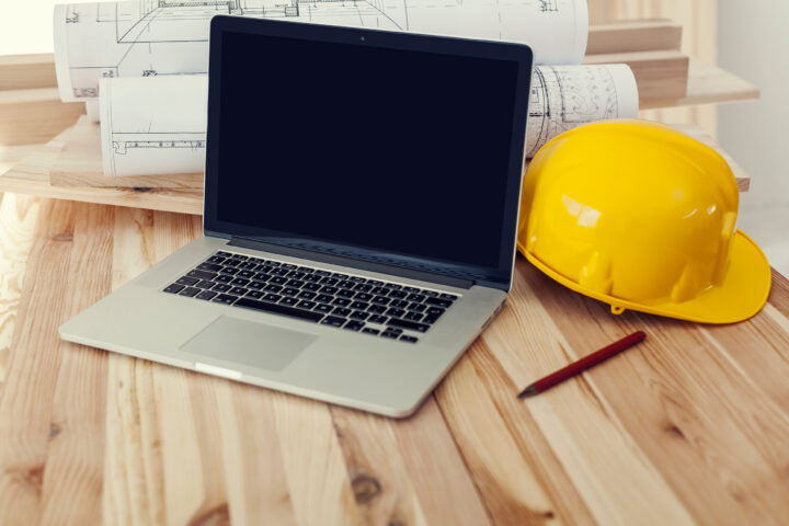 A laptop, yellow hardhat and set of blueprints sitting on a table.